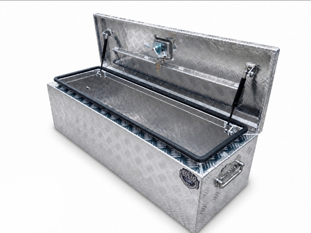 A silver diamond-plate metal storage trunk with its lid open, showing a black-lined interior compartment. The trunk features a lock, hinges, and a handle, photographed on a clean white background.
